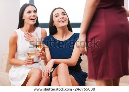 You are looking great! Two beautiful young women in evening gowns holding wine glasses and smiling while their friend standing in front of them and showing her dress