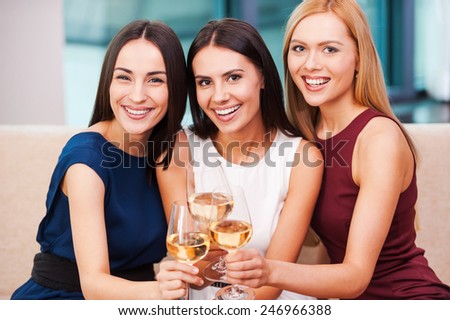 Enjoying great time together. Three beautiful young women in evening gown sitting on the couch and holding glasses with wine