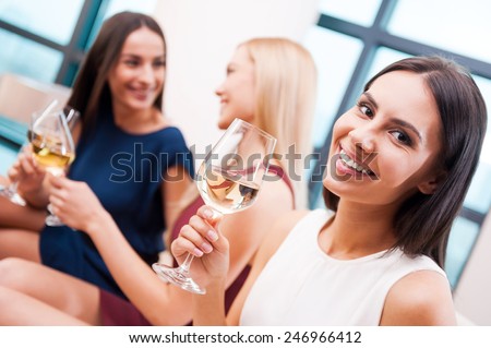 Enjoying good time with friends. Beautiful young woman in dress holding glass with white wine and smiling while two attractive women talking in the background