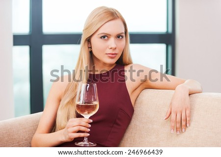 Sensual beauty. Beautiful young blond hair woman in evening gown sitting on the couch and holding glass with white wine