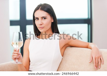 Enjoying white wine. Beautiful young blond hair woman in evening gown sitting on the couch and holding glass with white wine