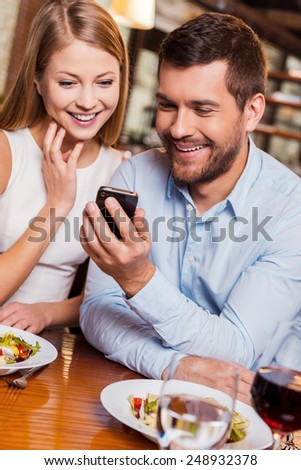 Just look at this! Beautiful young loving couple enjoying time in restaurant together while man showing something at his mobile phone and smiling