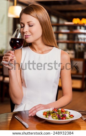 This wine perfect. Beautiful young woman holding glass with red wine and smelling it while sitting at the restaurant