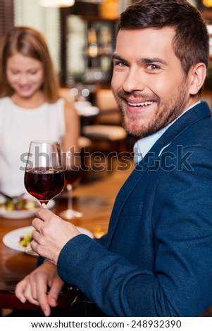 Spending great time in restaurant. Handsome young man holding glass with red wine and smiling while sitting at the restaurant with his girlfriend sitting in the background