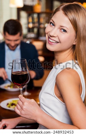 Enjoying time in restaurant. Beautiful young woman holding glass with red wine and smiling while sitting at the restaurant with her boyfriend sitting in the background