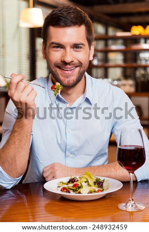 Enjoying meal at the restaurant. Handsome young man eating salad and smiling while sitting at the restaurant
