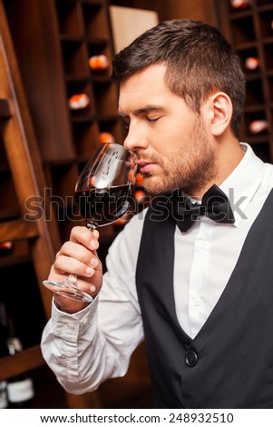 Testing wine. Confident male sommelier testing wine while standing near the shelf with wine