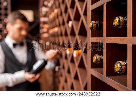 I recommend this wine. Confident male sommelier showing wine bottle and smiling while standing near the wine shelf