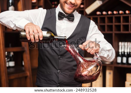 Pouring wine to decanter. Cropped image of confident male sommelier pouring red wine to the decanter while standing near the wine shelf