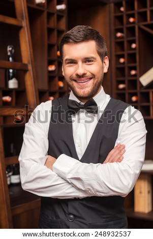 Confident sommelier. Confident male sommelier keeping arms crossed and smiling while standing near the wine shelf