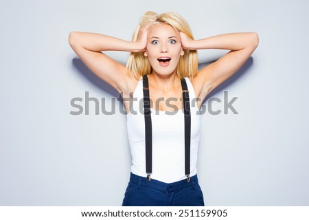 Shocked and happy. Surprised young woman holding hands in hair and staring at camera while standing against grey background