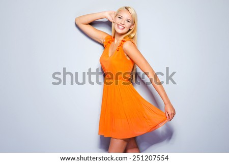 Spring is in my heart. Beautiful young woman in pretty dress holding hand behind head and smiling while standing against grey background
