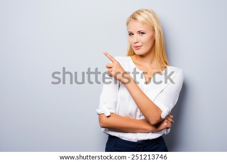 Over there! Beautiful young woman pointing away and looking at camera while standing against grey background