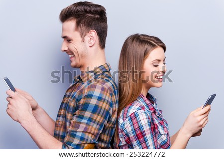 Romantic messages.  Beautiful young loving couple holding mobile phones and standing back to back against grey background