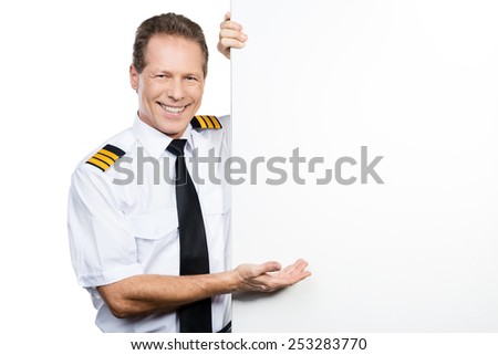 Can you believe it? Confident male pilot in uniform leaning at the copy space and pointing it with smile while standing against white background