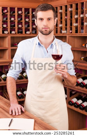 Confident winery owner. Confident young man in apron holding glass with red wine while standing in wine cellar