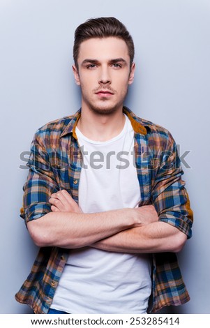 CHarming handsome. Handsome young man in shirt looking at camera and keeping arms crossed while standing against grey background