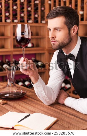 This wine is perfect. Confident male sommelier examining wine while looking at the wineglass and leaning at the wooden table with wine shelf in the background