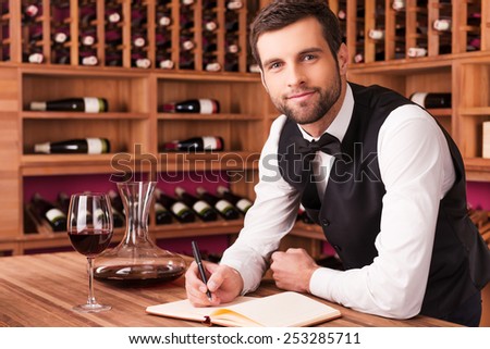 I know everything about wine. Confident male sommelier writing something in his note pad and looking at camera while leaning at the wooden table with wine shelf in the background