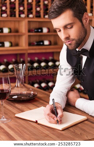 Making notes about wine. Confident male sommelier writing something in his note pad while leaning at the wooden table with wine shelf in the background