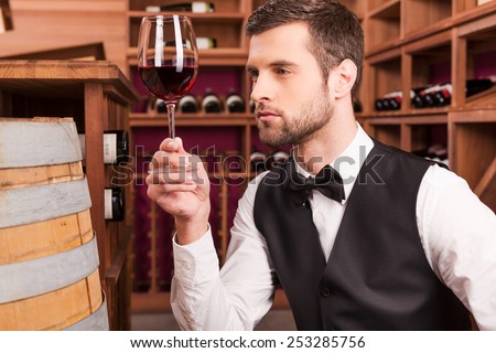 This wine is just perfect. Confident male sommelier examining wine while looking at the wineglass and sitting near the wine barrel