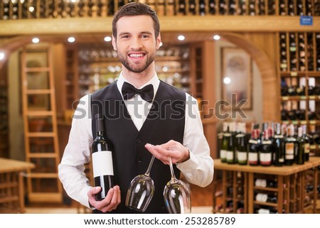 You should taste this perfect wine. Handsome young man in waistcoat and bow tie holding bottle and glasses while standing in liquor store