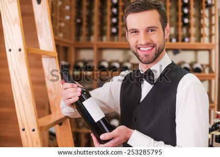 Choosing the best wine for you. Confident young man in waistcoat and bow tie holding bottle with wine and smiling while standing in liquor store