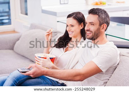 Enjoying their favorite show. Beautiful young loving couple bonding to each other and eating popcorn while sitting on the couch and watching TV