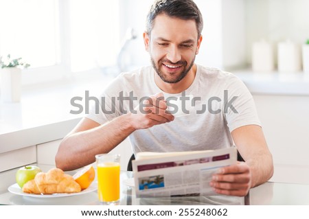 Starting day from good news. Handsome young man reading newspaper and smiling while drinking coffee and having breakfast in the kitchen