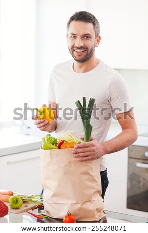 Using the freshest vegetables for my meal. Handsome young man unpacking shopping bag full of fresh vegetables while standing in the kitchen and smiling