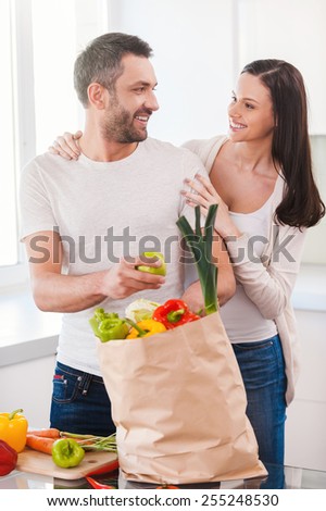 Enjoying happy and healthy life together. Beautiful young couple unpacking shopping bag full of fresh vegetables and smiling while standing in the kitchen together