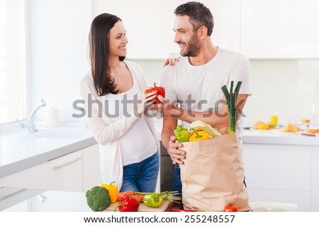 Living a healthy life together. Beautiful young couple unpacking shopping bag full of fresh vegetables and smiling while standing in the kitchen together