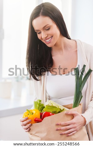Bag full of health and colors. Beautiful young woman unpacking shopping bag full of fresh vegetables and smiling while standing in the kitchen