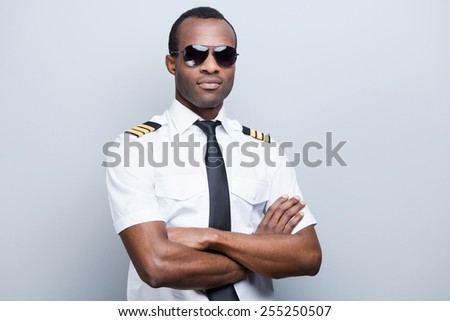 Confident and experienced pilot. Confident African pilot in uniform keeping arms crossed while standing against grey background