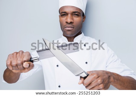 Ready to work. Confident young African chef in white uniform holding knifes in his hands and looking at camera while standing against grey background