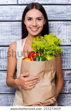Health in her hands. Beautiful young woman in apron holding paper shopping bag full of fresh vegetables and smiling while standing in front of wooden background