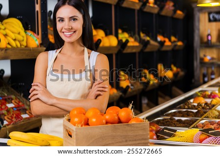 Welcome to fruit paradise! Beautiful young woman in apron keeping arms crossed and smiling while standing in grocery store with variety of fruits in the background