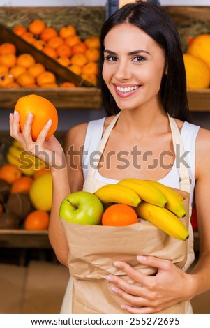 Join healthy lifestyle. Beautiful young woman in apron holding paper shopping bag with fruits and smiling while standing in grocery store with variety of fruits in the background