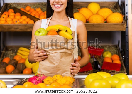 Selling health. Cropped image of beautiful young woman in apron holding paper shopping bag with fruits and smiling while standing in grocery store with variety of fruits in the background