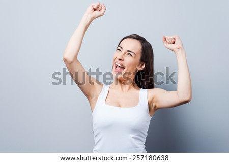 Can not hide her emotions. Beautiful young women keeping arms raised and smiling while standing against grey background