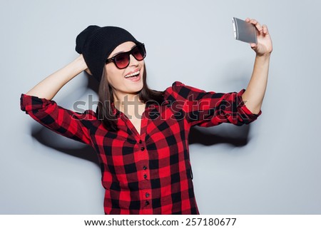 Saving memory of her new style. Portrait of beautiful young woman in glasses adjusting her hat while making selfie and standing against grey background