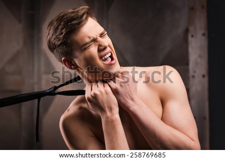 Got you! Frustrated young man choking by his necktie while standing against metal background