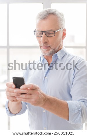 Typing business message. Serious grey hair senior man in shirt holding mobile phone and looking at it while standing in front of the window