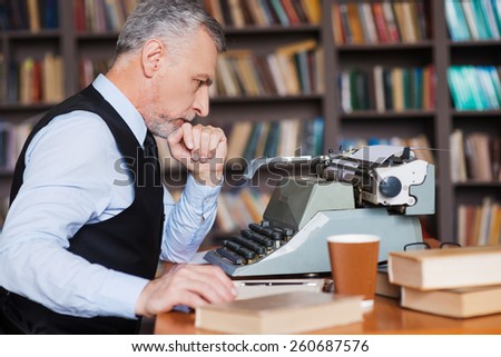 Waiting for inspiration. Side view of confident grey hair senior man in formalwear sitting at the typewriter and holding hand on chin with bookshelf in the background
