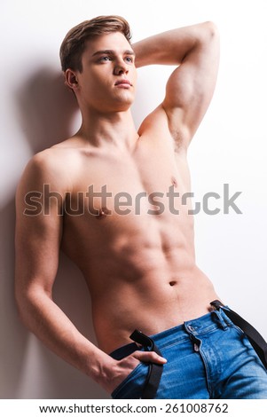 True masculinity. Handsome young muscular man holding hand in pocket and leaning the wall while standing against grey background