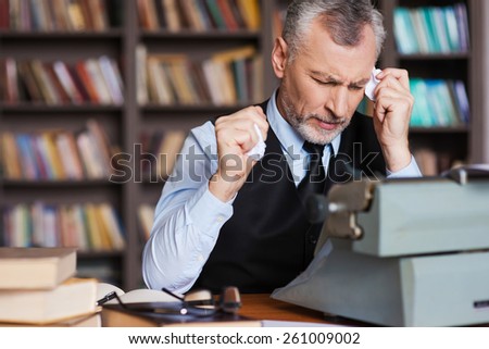 That is all wrong! Frustrated grey hair senior man in formalwear sitting at the typewriter and holding pieces of paper in his hands with bookshelf in the background