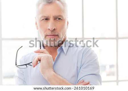 I think I have an idea. Thoughtful grey hair senior man in shirt holding his eyeglasses and looking away while standing in front of the window