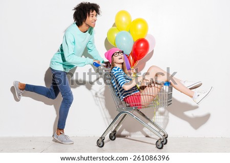 Spending great time together. Happy young man carrying his beautiful girlfriend in shopping cart and smiling while running against grey background
