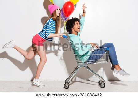 Spending great time together. Happy young women carrying his boyfriend in shopping cart and smiling while running against grey background