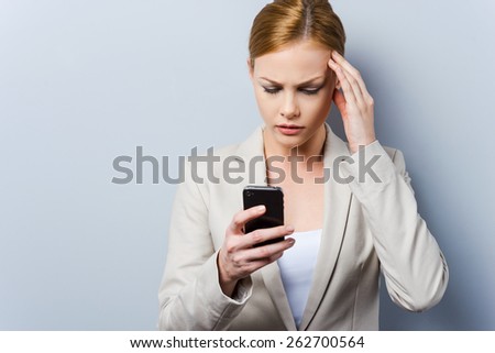 Having some troubles. Beautiful young businesswoman holding mobile phone and touching her face while standing against grey background